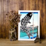 Infidu vintage wall art featuring a black koi fish hanging on a pole with intricate scales, set against a traditional Japanese landscape with a blue sky, serene water, and a classic bridge. displayed on the wall art in a white frame