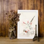 Vintage wall art with a bird surrounded by delicate flowers and plants in soft greens, reds, and whites, framed in white. displayed on the wall