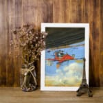 Infidu vintage wall art featuring a red airplane soaring through a bright blue sky with white clouds, evoking a sense of adventure and exploration. displayed on the wall art in a white frame