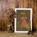 Infidu vintage wall art depicting a young girl in a pink dress holding a large green leaf over her head, set in a nostalgic outdoor scene with warm earthy tones. displayed on the wall art in a white frame