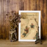 Vintage wall art of a nostalgic winter sledding scene with people in traditional winter clothing on a snowy slope, surrounded by trees. displayed on the wall in a white frame