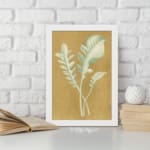 Vintage wall art of pale botanical fronds on an ochre background with a black frame. displayed on the wall in a white frame