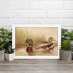 Infidu vintage wall art depicting two ducks swimming in a peaceful pond, surrounded by soft grass and gentle water waves, in calming green, brown, and beige tones. displayed on the wall art in a white frame