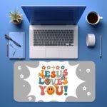 Light gray desk pad with “Jesus Loves You!” colorful text, surrounded by crosses, stars, hearts, smiley faces, and a happy emoji in vintage art style.